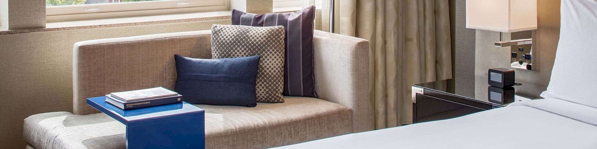 The image shows a modern hotel room with a bed, a lounge chair, pillows, a blue side table with books, and a window with a view of trees outside.