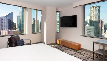 A modern room with large windows offering a cityscape view, a flat-screen TV, a chair, and a bed with white linens.
