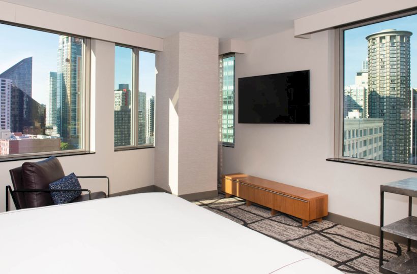 A modern room with large windows offering a cityscape view, a flat-screen TV, a chair, and a bed with white linens.