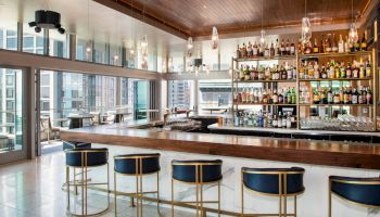 The image shows a modern bar with a wooden counter, blue barstools, and a well-stocked shelf of various drinks. Large windows offer a city view.