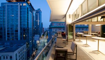 A modern balcony is shown with tables and chairs, overlooking a cityscape with tall buildings and a street visible below.