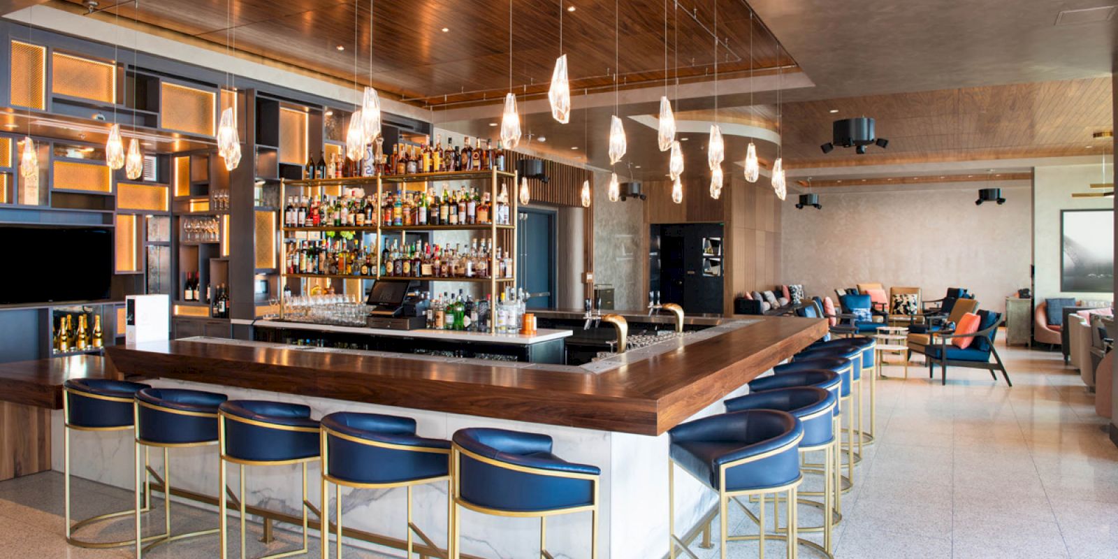The image shows a modern bar with a long wooden counter, blue chairs, hanging lights, a well-stocked shelf of bottles, and a chic seating area.