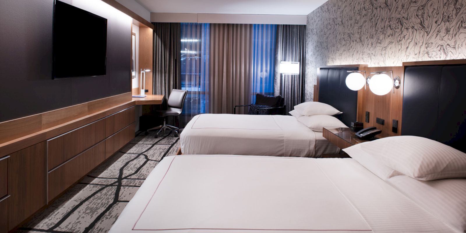 A modern hotel room with two neatly made beds, a wall-mounted TV, a work desk with a chair, and ambient lighting.