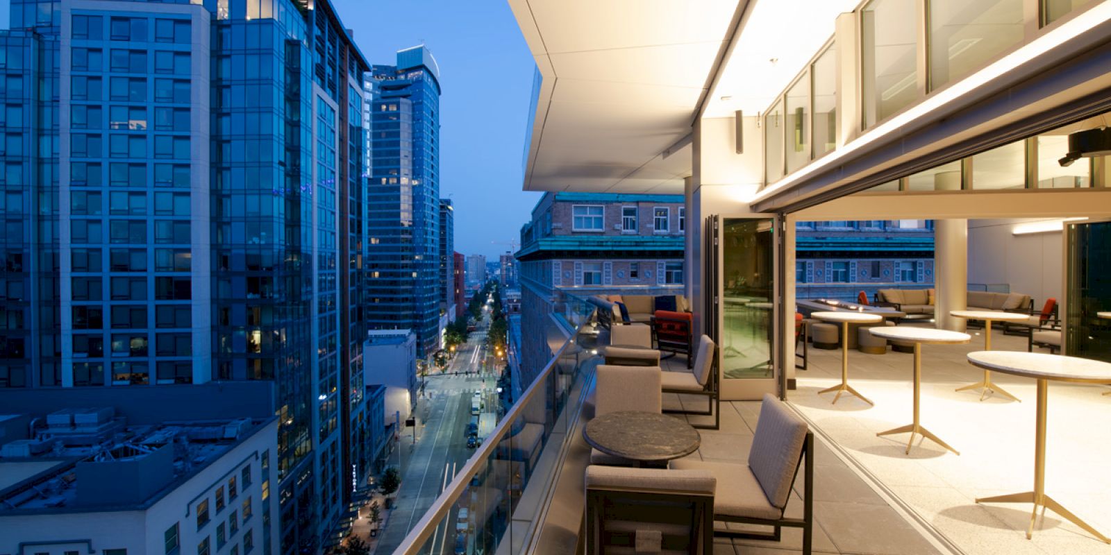 The image shows a modern cityscape with tall buildings at dusk and a spacious, furnished balcony of a high-rise building overlooking the street.