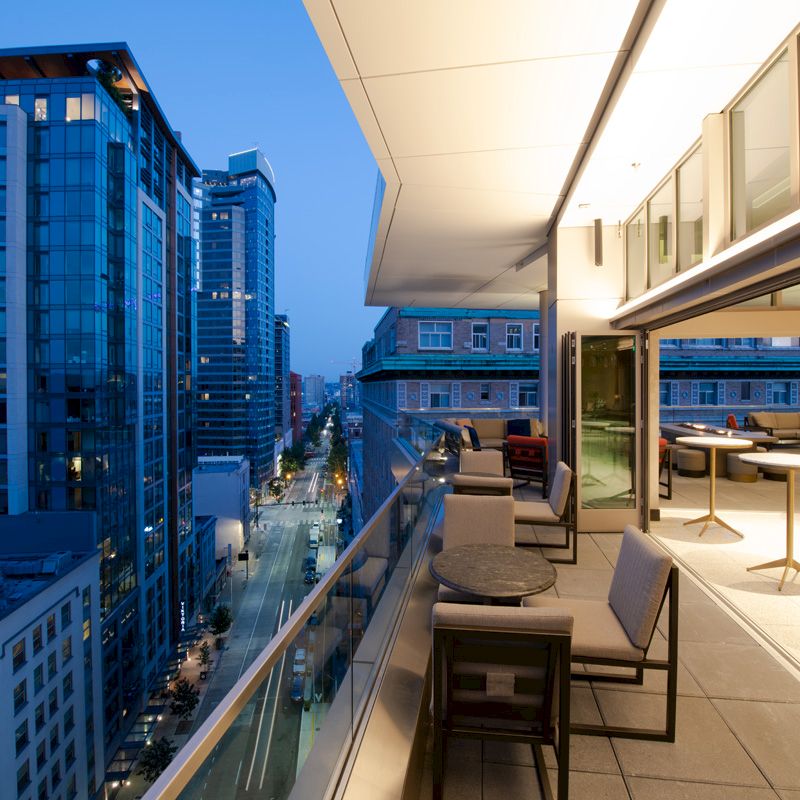 The image shows a modern cityscape at dusk with high-rise buildings and a spacious, stylish outdoor balcony with seating and tables.