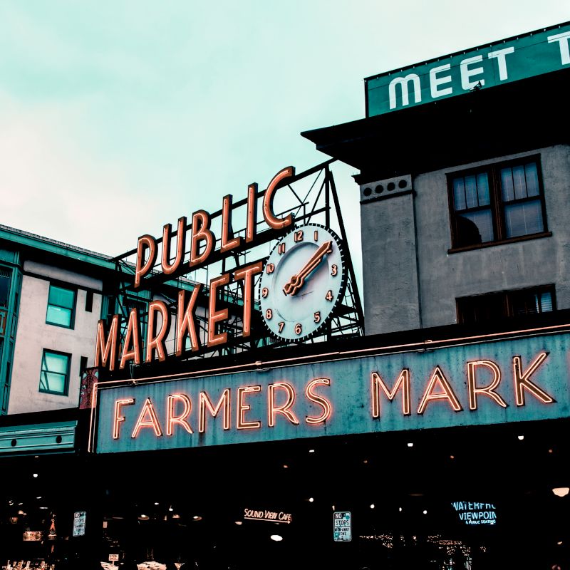 The image shows a public market with neon signs for "Public Market" and "Farmers Market," featuring a clock and older style buildings.