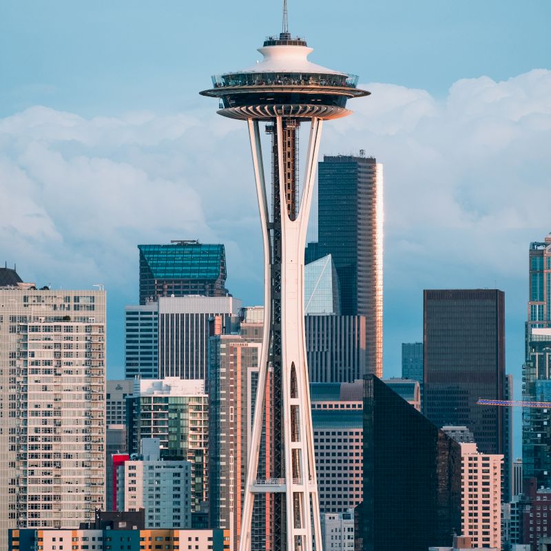 The image features the iconic Space Needle in Seattle, surrounded by various modern skyscrapers and buildings, with a cloudy sky in the background.