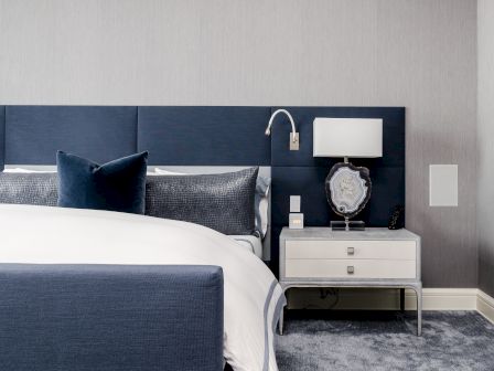 A modern bedroom has a bed with blue and white bedding, a navy headboard, and a nightstand with a lamp and decorative object.
