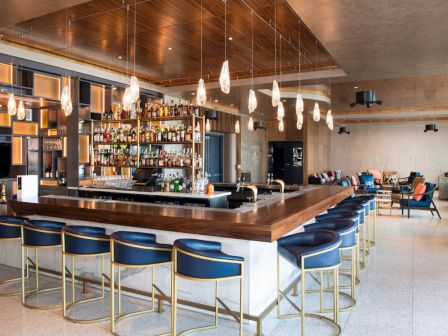 A modern bar with blue stools, a wooden countertop, and hanging lights, with shelves stocked with various bottles behind it.