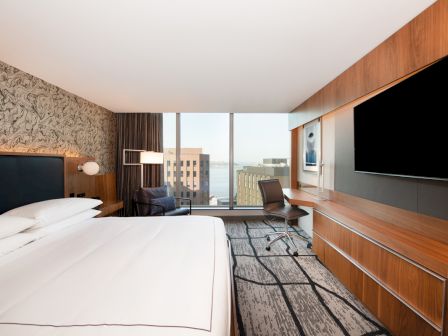 A modern hotel room with a large bed, desk, chair, and a wall-mounted TV. The window offers a view of city buildings and a bit of water.