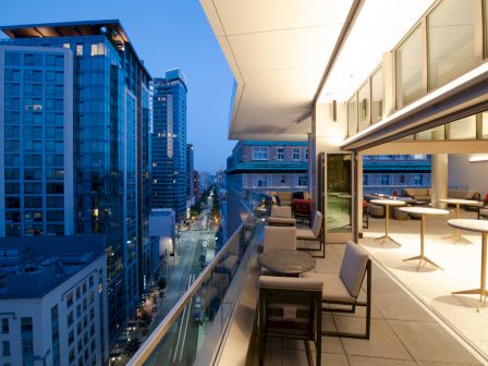 A modern balcony overlooks a bustling city street with tall buildings. The balcony features seating and tables, with a well-lit indoor area adjacent.