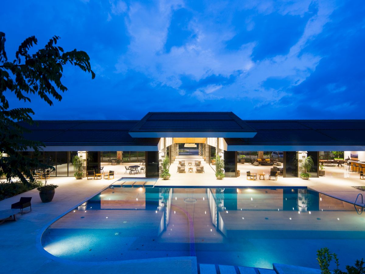 A luxurious modern building is shown with an illuminated outdoor swimming pool at dusk, surrounded by seating areas and greenery.