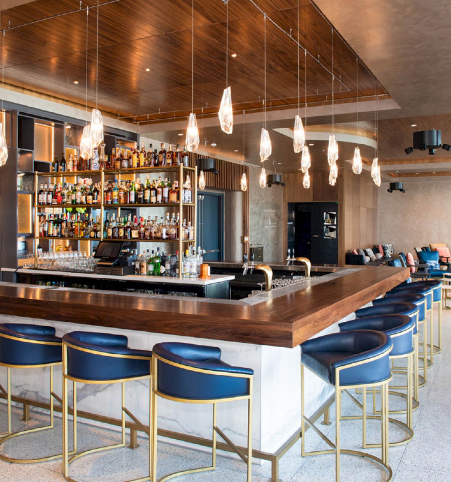 A modern bar with a wooden counter, blue chairs, hanging lights, and a fully stocked shelf backdrop; cozy seating area in the background.