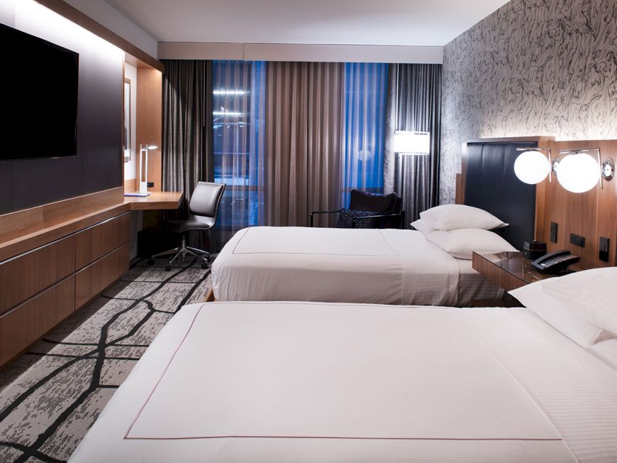 This image shows a modern hotel room with two beds, a desk with a chair, a large TV, and floor-to-ceiling windows with curtains.