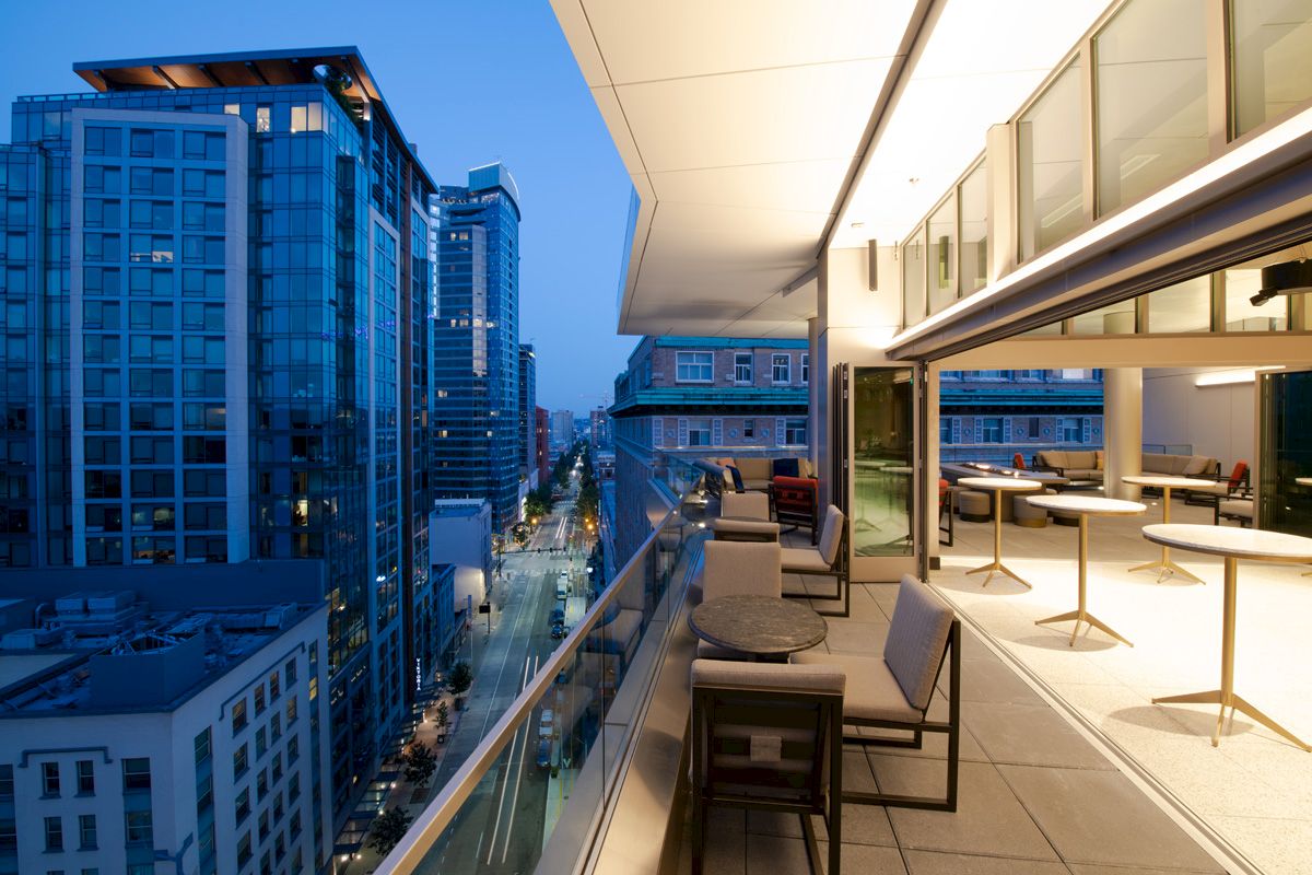 A modern balcony with seating overlooks a city street with tall buildings. The interior is stylish with tables and chairs. Evening time.