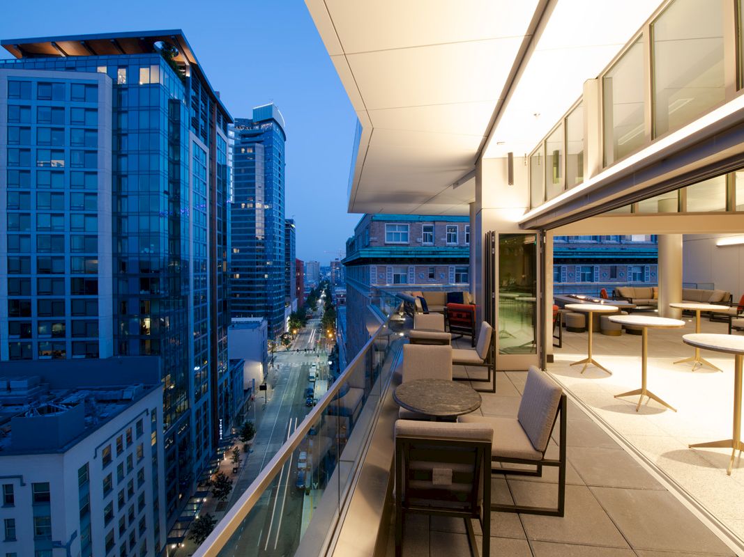 A modern balcony with seating overlooks a city street with tall buildings. The interior is stylish with tables and chairs. Evening time.