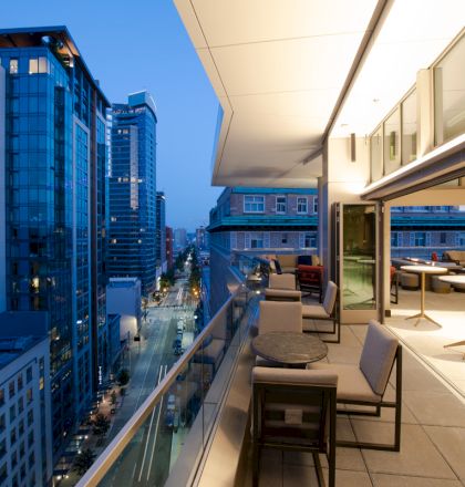 A modern cityscape at dusk with high-rise buildings and a spacious balcony featuring tables and chairs on the right side.