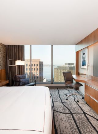 A modern hotel room features a large bed, a wall-mounted TV, a desk with a chair, a cozy seating area, and a window offering a city view.