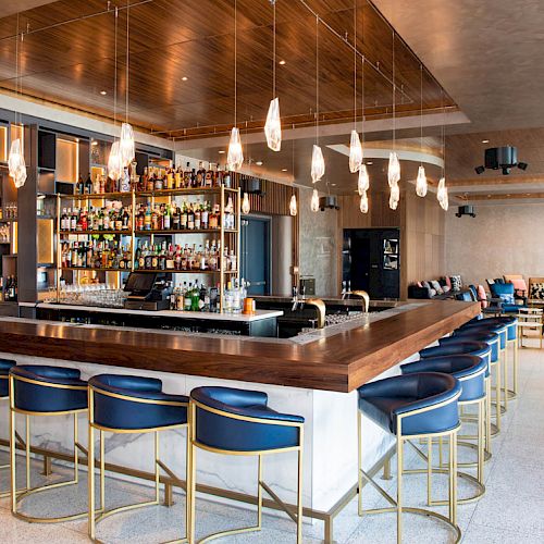 The image shows a modern bar with a wooden counter, blue chairs, hanging pendant lights, and a fully stocked bar in the background.