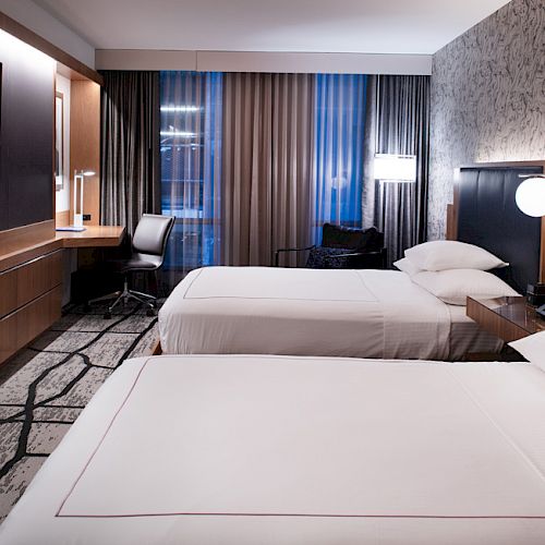 The image shows a modern hotel room with two neatly made beds, a large TV, a desk with a chair, and contemporary lighting.