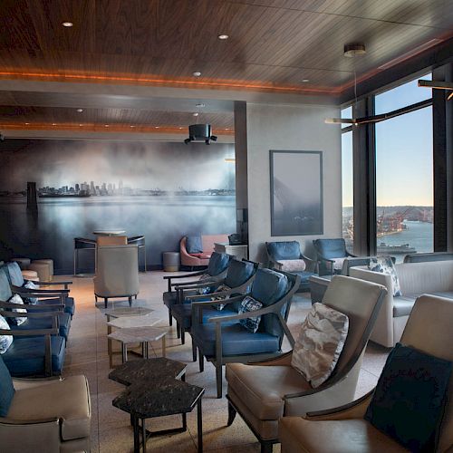 A modern lounge with numerous seats, a mural of a city skyline, and large windows offering scenic views of a waterfront and a Ferris wheel.