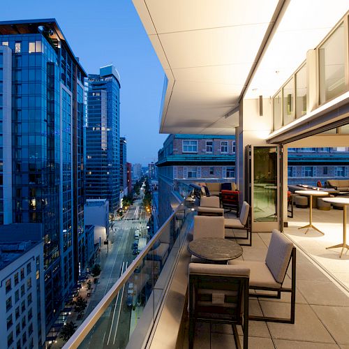 A modern cityscape featuring high-rise buildings and a scenic balcony with stylish furniture, overlooking a busy street at dusk.
