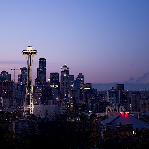 The image shows the Seattle skyline at dusk, featuring the Space Needle prominently in the foreground with Mount Rainier in the background.