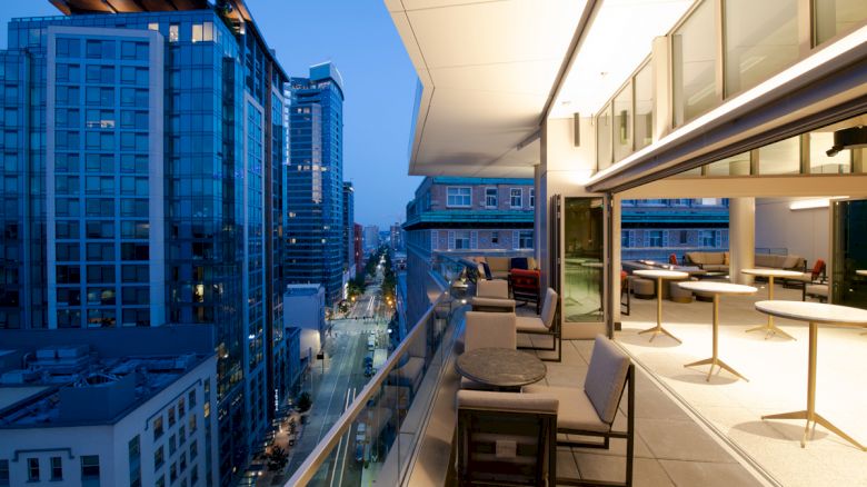 A modern cityscape at dusk, with high-rise buildings and a luxurious rooftop patio featuring furniture and lighting, overlooking the street below.