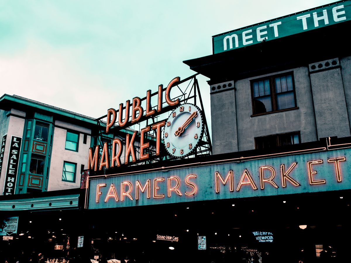 The image shows an iconic sign for a public farmers market, with a clock and neon lights, against the backdrop of historic buildings.