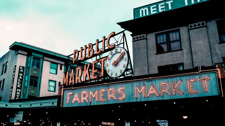 The image shows an iconic sign for a public farmers market, with a clock and neon lights, against the backdrop of historic buildings.