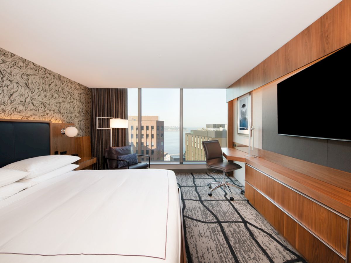 A modern hotel room with a large bed, a flat-screen TV, a desk, a chair, and a window offering a city view, including a glimpse of the water.