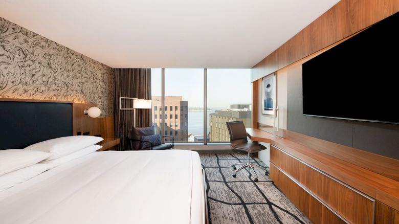 A modern hotel room with a large bed, a flat-screen TV, a desk, a chair, and a window offering a city view, including a glimpse of the water.