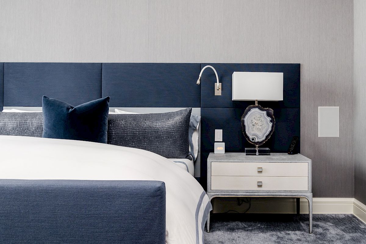 A modern bedroom features a navy blue headboard, white bed linens, a nightstand with a lamp, and minimalist decor.