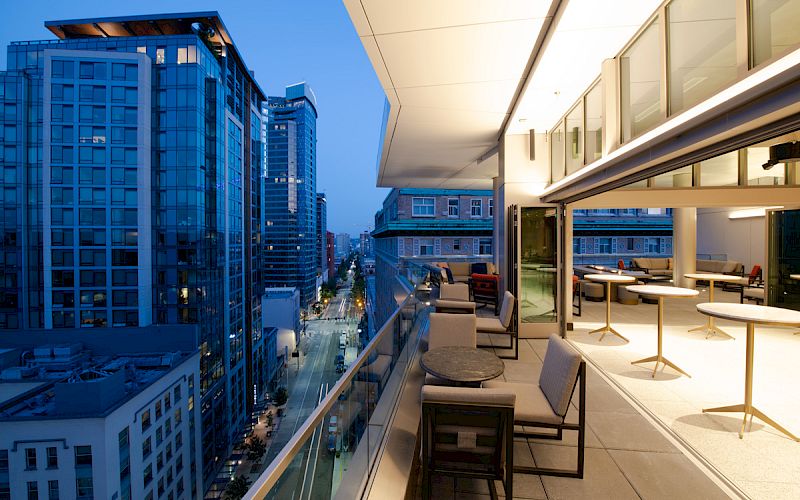 A modern cityscape at dusk viewed from a high balcony with outdoor seating, tables, and a well-lit indoor area.