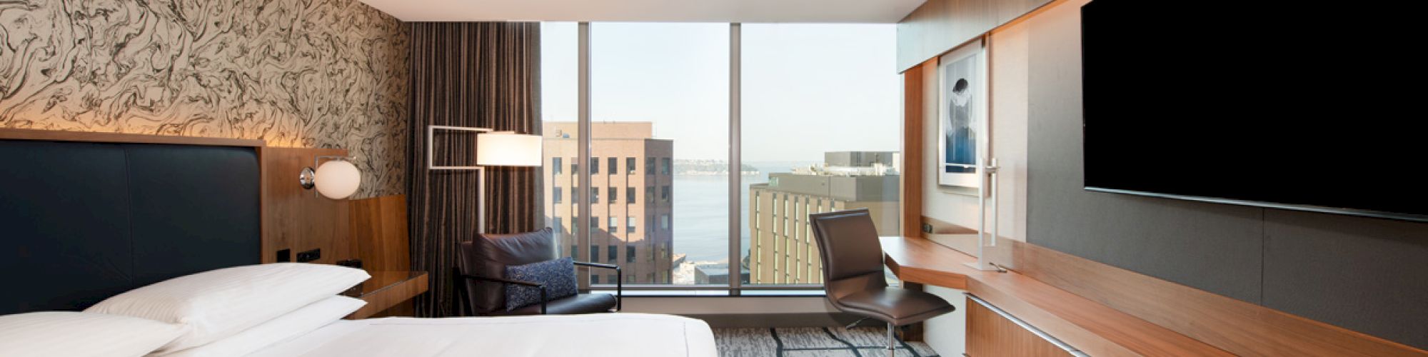 A modern hotel room with a large bed, wall-mounted TV, desk, chair, and a window offering a cityscape view with buildings in the background.